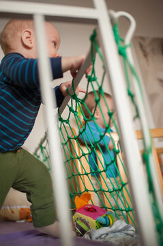 Sweet twin baby brothers standing in their cozy wooden crib during the bright afternoon to reach for toys through the soft green safety netting