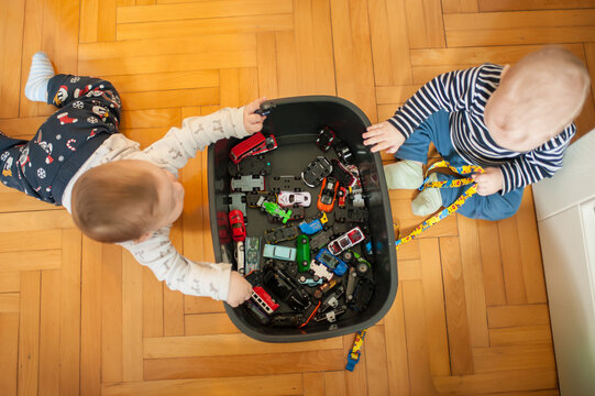 Toddlers crawl on the hardwood floor, reaching into a black container to discover and play with a variety of miniature toy cars