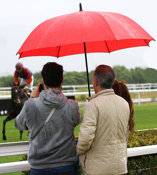 P&uacute;blico con paraguas rojo en hip&oacute;dromo, espectador fotografiando con tel&eacute;fono m&oacute;vil y grabando caballo y jockey de carreras en plan de ocio en familia d&iacute;a de lluvia racecourse an red umbrella