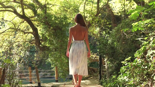 A woman in a backless dress walks along a sunlit forest path