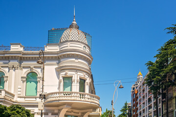 Upper part of corner white building facade with dome, spire, arched windows, round balcony with balustrade against city buildings, blue sky. © Elena