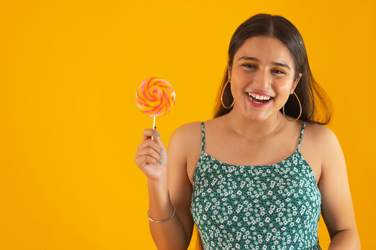 Closeup portrait of a cheerful teenage girl holding lollipop against yellow background