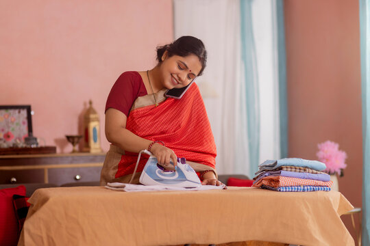 Portrait of a happy woman talking on mobile phone while ironing clothes at home