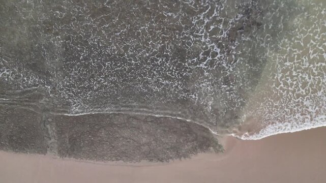 Aerial drone footage of foamy white waves moving toward the beach, colliding with dark coastal rocks. Captures the raw beauty of sea, surf, and shoreline in motion.