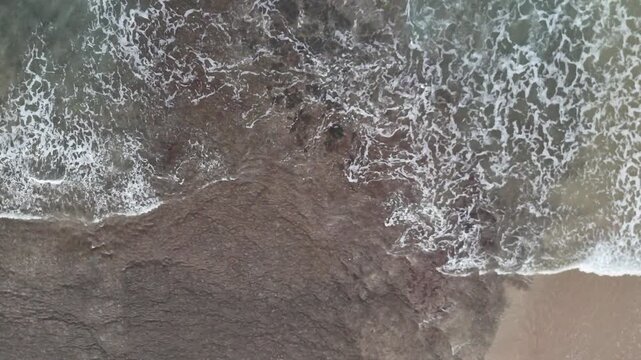 Aerial drone footage of foamy white waves moving toward the beach, colliding with dark coastal rocks. Captures the raw beauty of sea, surf, and shoreline in motion.
