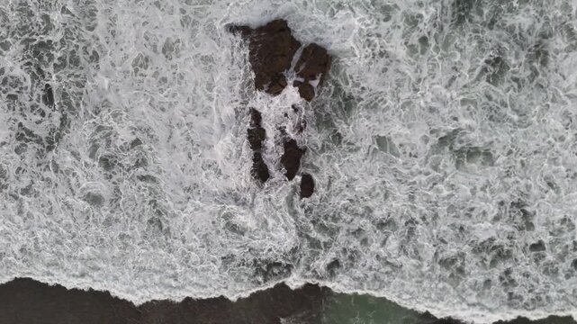 Aerial drone footage of foamy white waves moving toward the beach, colliding with dark coastal rocks. Captures the raw beauty of sea, surf, and shoreline in motion.