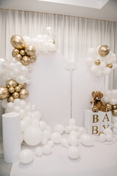 Photo zone for a birthday, baptism, or baby shower featuring white arched panels decorated with white and gold balloon garlands, a white pedestal, and a plush teddy bear on "BABY" letter blocks.