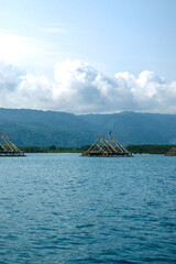 Traditional floating fish traps resting on calm blue water with distant green hills beneath a bright cloudy tropical sky. © tomz