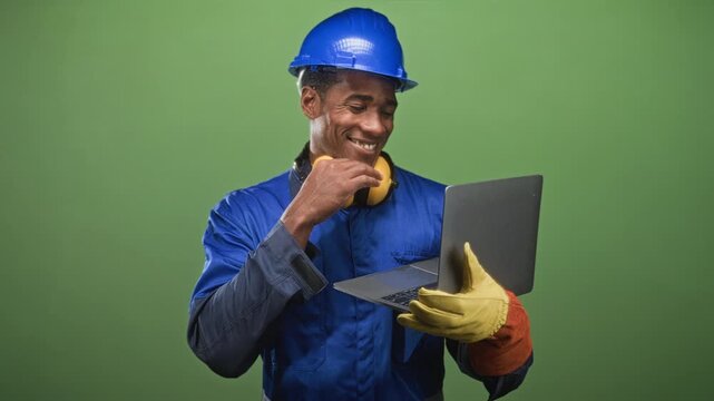 Man builder in blue hardhat and gloves holding laptop and adjusting yellow earmuffs while smiling in studio; confidence productivity safety.