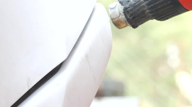 Mechanic uses heat gun to remove car bumper scratch. Auto body repair process of fixing paint damage with heating tool. Automotive maintenance and refurbishment.