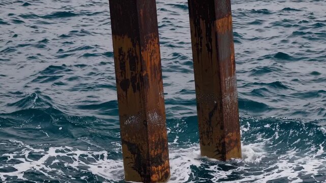 Rusty wooden pilings submerged in ocean water create splashes as waves crash against them, showcasing the dynamic interaction between structure and sea