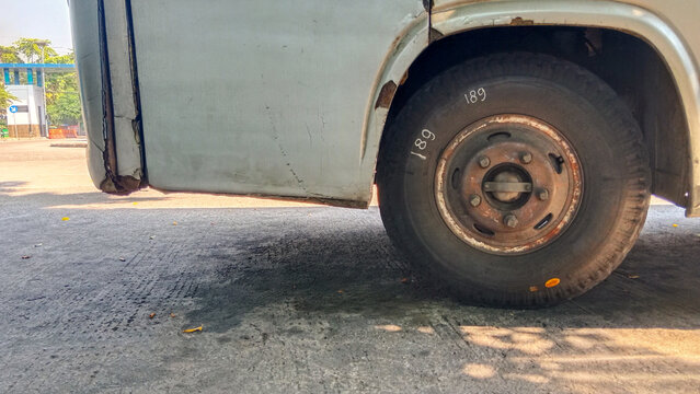 Close up zoomed of a white bus wheel and fender parked on asphalt. The vehicle has a shiny hubcap and tire tread details visible under sunlight.