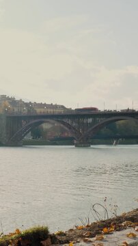 Vertical video captures a bus traveling across Maribor Bridge over the Drava River, scenic autumn foliage and soft daylight in Slovenia, ideal for social media.