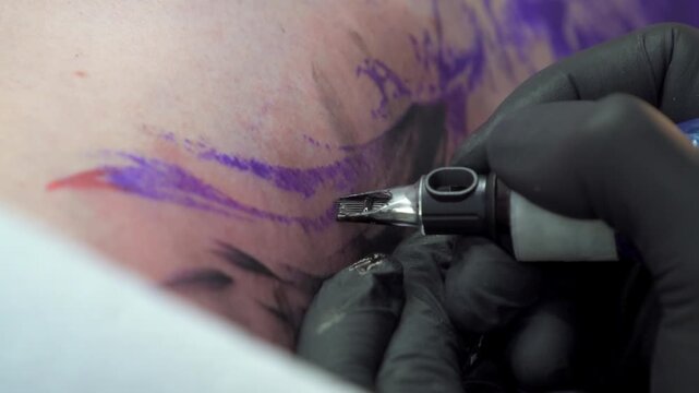 Detailed close-up shot of a tattoo artist with black gloves using a machine to ink a black and purple tattoo on human skin in a studio setting.