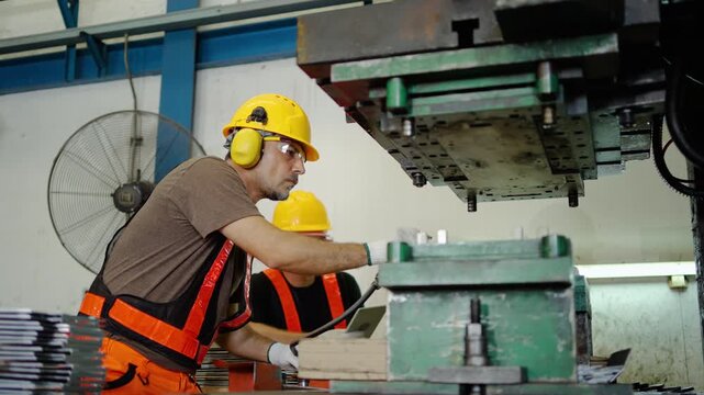 Professional male factory worker in protective safety gear uses a pneumatic air gun to clean heavy industrial stamping press machinery. Modern manufacturing plant emphasizing teamwork and production.