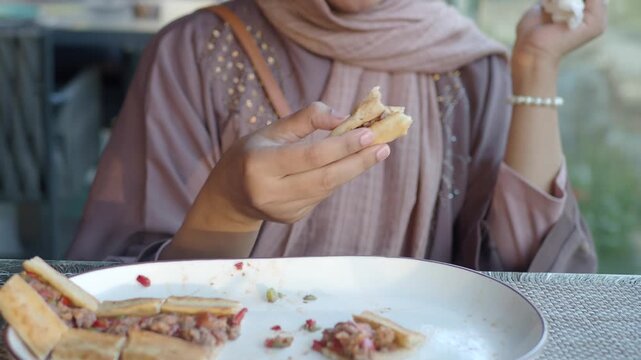 Young woman in hijab enjoys delicious breakfast at outdoor cafe, close-up shot of her hands holding and eating a piece of pita bread with meat and vegetables