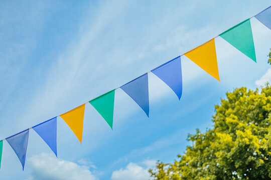 Blue, green, yellow, and white triangular pennant flags stretch across a bright sky above leafy trees, with ample copy space and a cheerful outdoor atmosphere