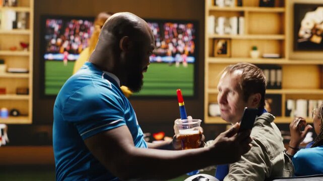 Man on apartment couch holding smartphone while following football broadcast, telling friends about negative online match results while soccer supporters drink beer at home, frustrated by match.