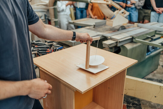 Craftsperson demonstrates a handmade wooden balance model on a cabinet while visitors observe inside a woodworking shop with machinery in the background