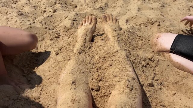 Kids burying and covering their feet with warm wet sand on a sea beach, summer family vacation and childhood fun concept