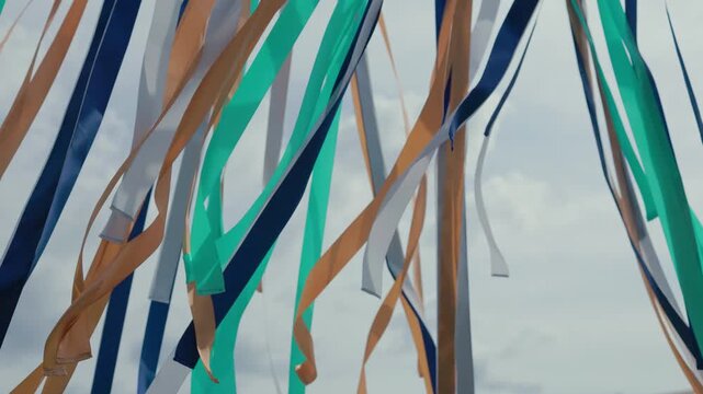 Orange, green, blue, and white fabric ribbons hang and flutter beneath an outdoor frame against a bright cloudy sky, creating a lively festive pattern