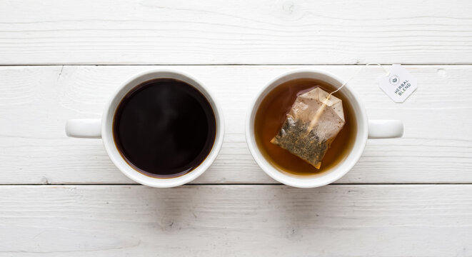 Two white ceramic mugs sit side by side on a rustic white wooden tabletop containing dark roasted hot coffee and a soothing herbal tea blend with a paper tea tag
