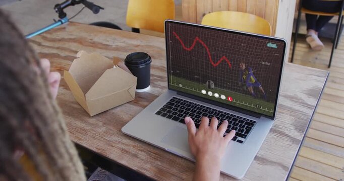 Male typing, handling takeout pastry while red plot animating on laptop to monitor market at cafe