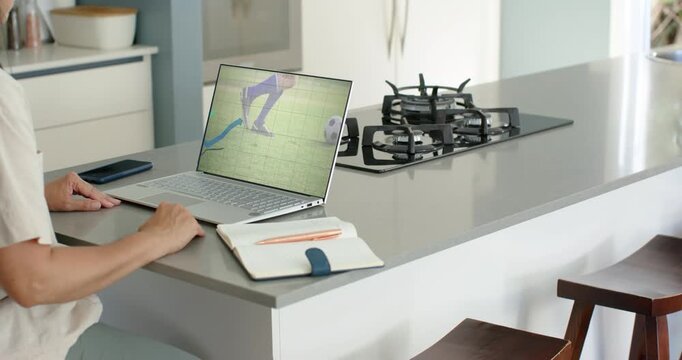 Mature woman watching soccer clip on laptop, resting hands and leaning at kitchen island, notebook
