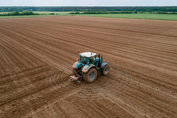 Fototapeta premium Agricultural tractor moves across vast brown soil. Circles mark its path as it plows the field. Farmland stretches wide with green trees in distance. No people appear, no animals present in the scene