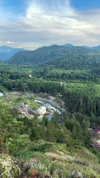The historic Chemal hydroelectric power station building stands along a rushing river channel surrounded by dense green pine forests and rolling mountain slopes under a scenic cloudy sky, 4k