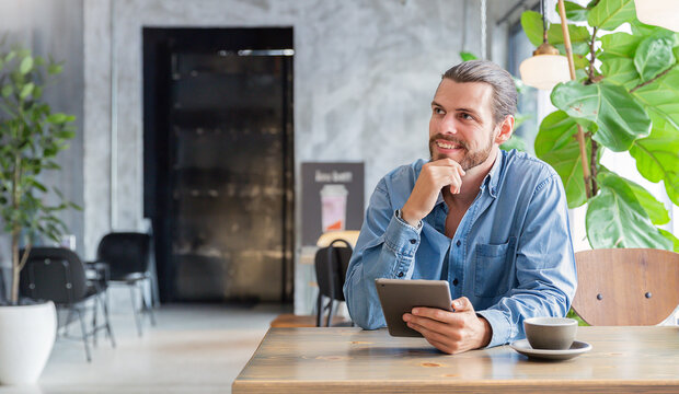 Young tattooed man sitting at a modern cafe, using a tablet near a window with coffee and a small cactus nearby. Relax slow life with technology