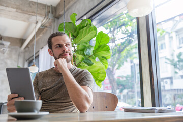 Fototapeta premium Young tattooed man sitting at a modern cafe, using a tablet near a window with coffee and a small cactus nearby. Relax slow life with technology