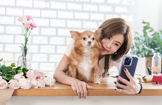 Portrait of smile asian woman taking selfie with small dog on desk surrounded by flowers in bright floral studio workspace Asia girl playing with little dog. Love together family.