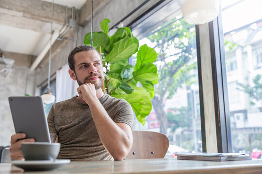 Young tattooed man sitting at a modern cafe, using a tablet near a window with coffee and a small cactus nearby. Relax slow life with technology