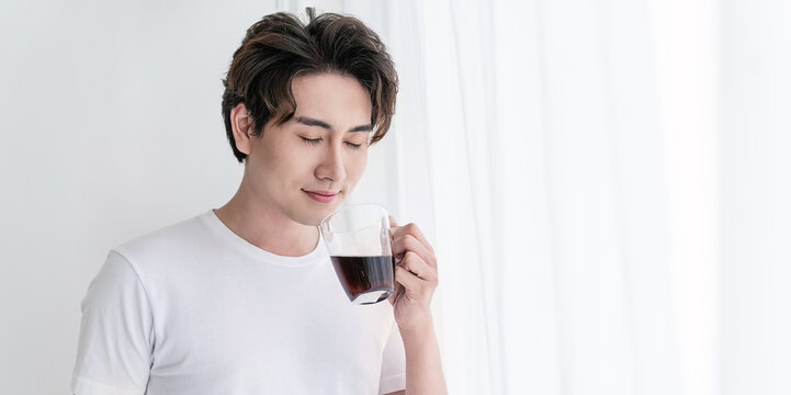 Portrait of young handsome asian man hands holding coffee glass cup morning spring time in white bedroom. Happy cheerful relaxing in summer. Korean male guy wakeup, university asia lifestyle concept