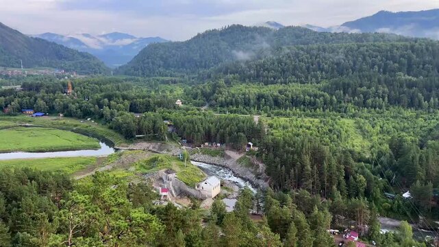The scenic panorama of the Chemal hydroelectric power station on a winding river surrounded by vast pine forests, mountains with mist on their peaks, and the wide Katun River flowing in the distance u
