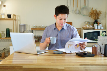 Fototapeta premium Young Asian professional reviews business charts and reports while drinking coffee at home kitchen, ideal for remote work and freelance concepts.