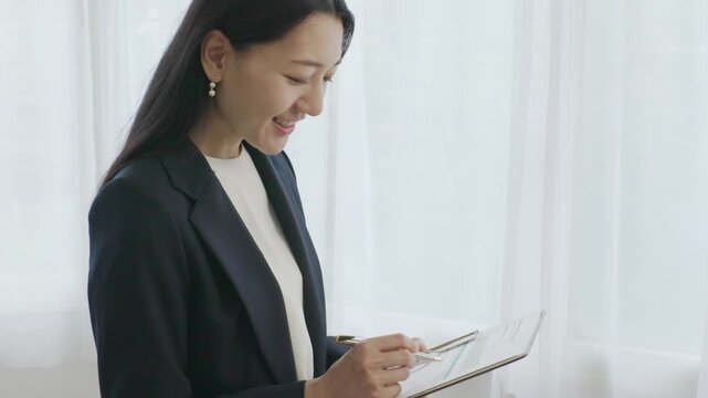 Young Japanese businesswoman taking notes on binder smiling by window