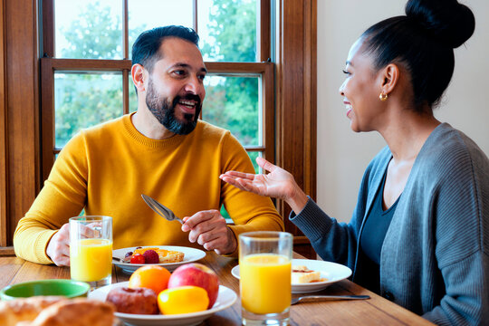Young adult couple sharing breakfast, talking and smiling across table at home, expressing connection and daily routine. Ideal for family lifestyle, relationship, communication campaigns