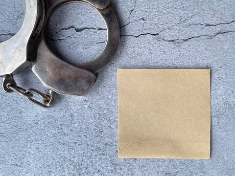 Blank sticky note beside metal handcuffs on textured concrete background for legal, crime prevention and awareness concepts.