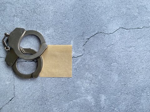 Metal handcuffs with blank sticky note on cracked concrete background representing crime, justice, legal awareness and copy space.