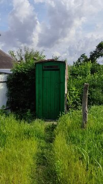 Old green wooden outdoor toilet surrounded by tall grass and lush greenery, vertical