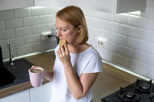 Woman holding pink mug and eating cookie in kitchen. Concept of snack time and morning coffee.