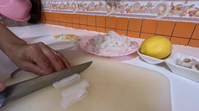 Fresh squid being sliced into strips on a cutting board for cooking. Preparation highlights tender texture, with lemon and garlic nearby for seasoning, commonly used in seafood dishes.