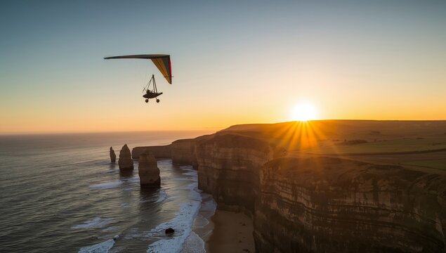 Hang glider soaring over coastal cliffs at sunset, with ocean waves crashing against the shore and the sun setting on the horizon