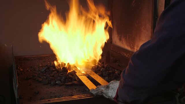 Close-up of a skilled blacksmith placing a steel bar into a traditional coal forge as intense flames surround the hot metal during the forging process.
