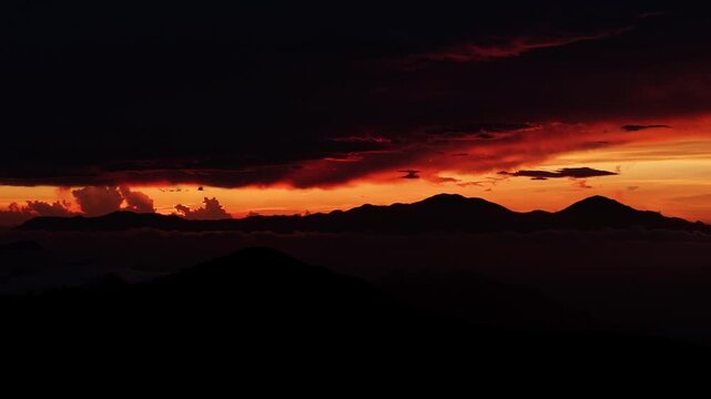 Dramatic orange sky, glowing horizon, dark mountain silhouettes, and storm clouds create a spectacular natural scene.
