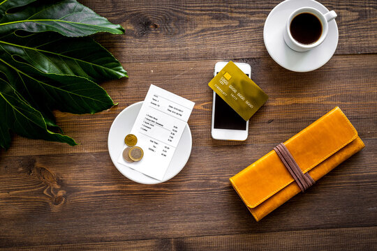 Pay restaurant bill. Bill, bank card, wallet, coins near cup of coffee on dark wooden table top view
