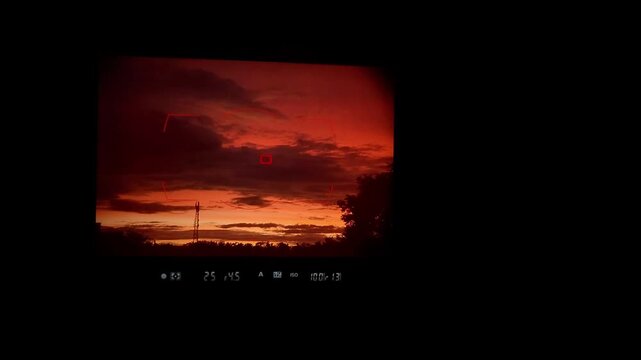 DSLR camera viewfinder display looking at a dramatic and vibrant red sunset with dark clouds over a silhouetted landscape.