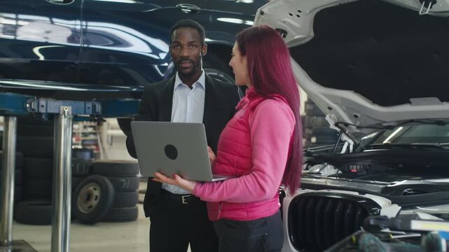 Woman mechanic showing a vehicle diagnostic result on a laptop to a male customer in a workshop with lifted cars and professional equipment.